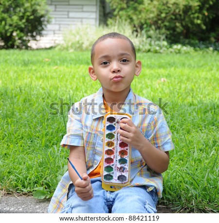 Little Boy Puckering Lips looking at camera holding paint set sitting on grass