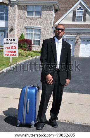 Young Businessman with suitcase standing in front of home relocation for sale realtor sign on front yard lawn of brick suburban home