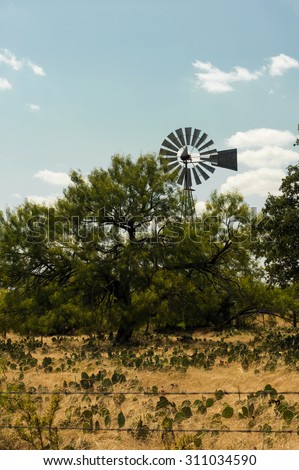 Country Windmill in West Texas - Prickly Pear cactus and sun dried weeds with Mesquite and Live Oak Trees in the foreground.  Blue sky with scattered clouds in the background.