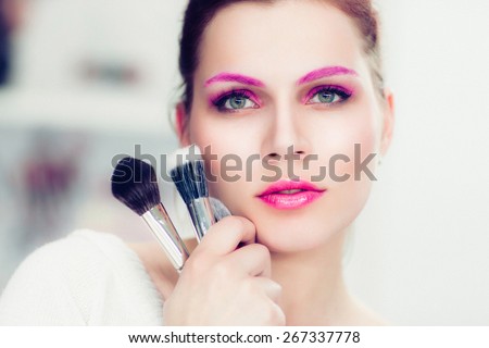 The makeup artist with bright pink make-up holds powder brushes. She touches with them a cheek. Studio portrait.