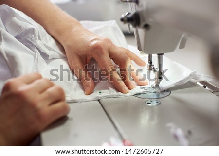 Close up side top view of cropped female hands sewing white fabric on professional manufacturing machine at seamstress workplace. Tailor hands holding textile for dress production. Blurred background Foto stock © 