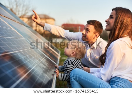 Man shows his family the solar panels on the plot near the house during a warm day. Young woman with a kid and a man in the sun rays look at the solar panels. Stock fotó © 