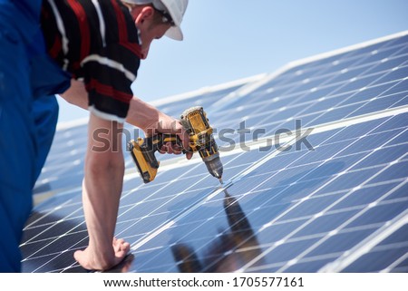 Male engineer in protective helmet installing solar photovoltaic panel system using screwdriver. Electrician mounting blue solar module on roof of modern house. Alternative energy ecological concept.