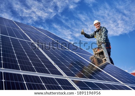 Low angle view portrait of a smiling worker, installing solar batteries, who is standing on ladder at solar plant against blue sky, showing thumb up. Concept of alternative sources of energy.