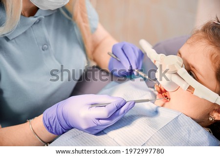 Female dentist checking child teeth with dental explorer and mirror while girl lying in chair with inhalation sedation at dental office. Concept of pediatric, sedation dentistry and dental care. Stock foto © 