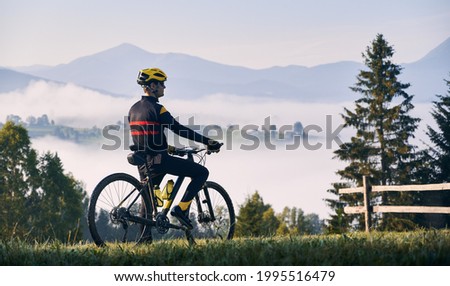 Cyclist standing with bicycle on grassy hill and looking at beautiful misty mountains. Male bicyclist enjoying the view of majestic mountains during bicycle ride. Concept of sport, bicycling, nature.