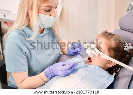 Female dentist checking child teeth with dental explorer and mirror while girl lying in dental chair with inhalation sedation at dental office. Concept of pediatric, sedation dentistry and dental care Stock foto © 