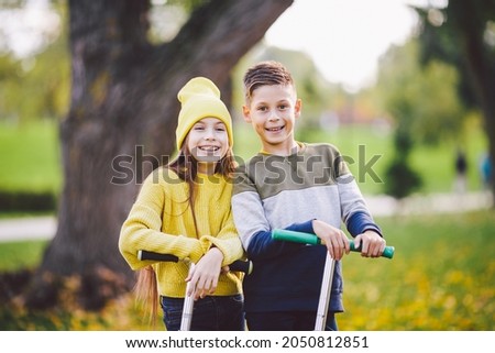 Caucasian children twins boy and girl schoolchildren age pose together in sunny weather in autumn park after riding on kick scooters. Happy kids riding kick scooters in public park. Kids sport.