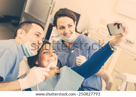 Happy Patient, Dentist and Assistant Taking Selfie All Together. Patient is Holding Smart Phone, Dentist and Assistant are Holding their tools. Focus on Patient Eyes