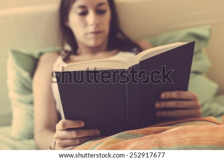 Young Woman Reading a Book on the Bed. The bed has green and orange Sheets. The Girl is alone, she is Holding the Book with both Hands while Lying down on the Bed. Focus on the Book.