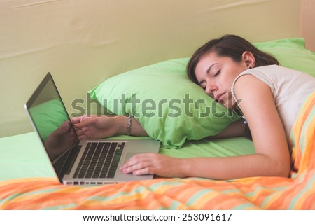 Young Woman Falling Asleep while Using Notebook on Bed. She is Alone. The bed has green and orange Sheets. She still Have her Hands on Keyboard. Technology or Internet addiction theme.