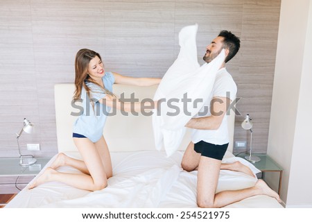 Happy Couple Having Pillow Fight in Hotel Room. They Wear Underwear and stand on bendend Knees on the Bed. Everyone is Holding a Pillow. The Woman has Long Blonde Hair, the Man has Black Short Hair.