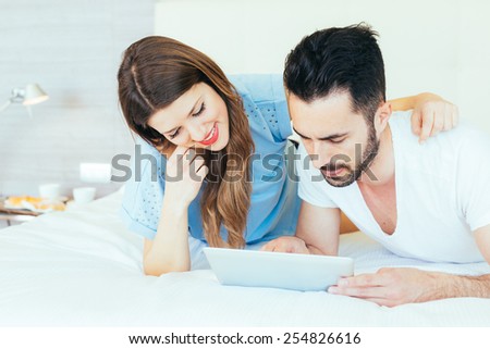 Young Couple with Digital Tablet on Bed. They are in a Modern Hotel Room and They wear Underwear. The Man is holding the Tablet with his left hand. Technology addiction and love concepts.