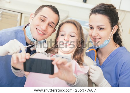 Happy patient, dentist and assistant taking selfie all together. Patient is holding smart phone, dentist and assistant are holding their tools. Focus on patient eyes