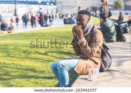 Young man having lunch break in London next to Tower Bridge. He is seated on a concrete low wall and is eating a sandwich.
