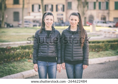 Female twins portrait at park. They wear black jackets and jeans. Buildings and cars on background. They stand side by side.