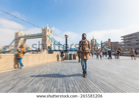 Man walking in London on Thames sidewalk, with Tower Bridge and blurred people on background. He is looking away. Photo taken on a sunny winter day.