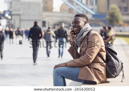 Black man talking on mobile phone in London. He is seated on a concrete bench, on background there are a lot of blurred persons and the Tower Bridge