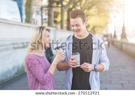 Young couple walking in London, talking and smiling, and holding a cup of tea. They are in their twenties, wearing light clothing and holding hands.Backlight flare added.