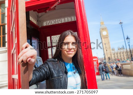 Beautiful young woman in London with Big Ben and red phone booth on background. She is holding the door open and looking at camera. Focus on the face.