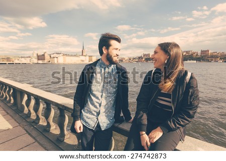 Young hipster couple visiting Stockholm. They are walking with sea and old town on background. Both are wearing sunglasses and a black jacket.