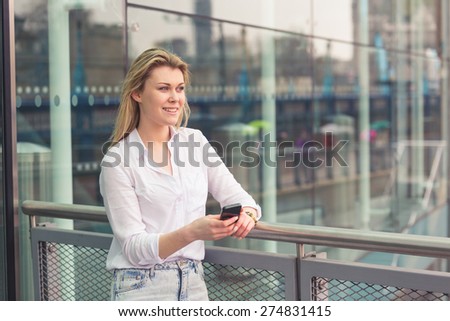 Young woman typing on smart phone in front of a modern glazed building in London. She is blonde, wearing a white shirt and staying by three quarters.
