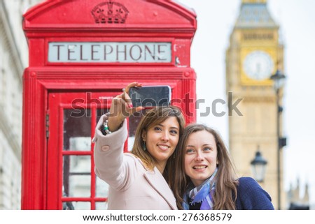 Two beautiful women taking a selfie in London with Big Ben and red phone booth on background. They are in their twenties, holding the phone and looking at it. Focus on the face.