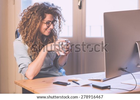 Young woman working at home or in a small office, vintage hipster clothing, curly hair. Cup of tea or coffee on the desk with some technological devices.