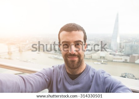 Young man taking a selfie with London cityscape on background. He is holding a smart phone and looking at it smiling.