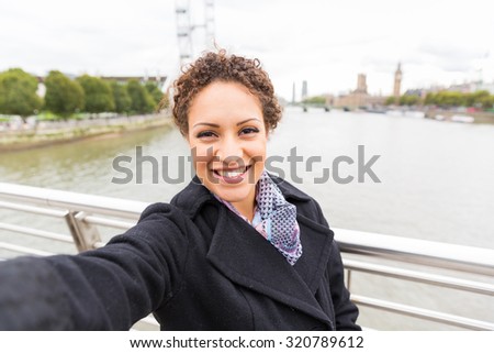 Beautiful mixed race woman taking a selfie in London with Thames river and Big Ben on background. She is holding the camera with one hand and looking at it smiling.