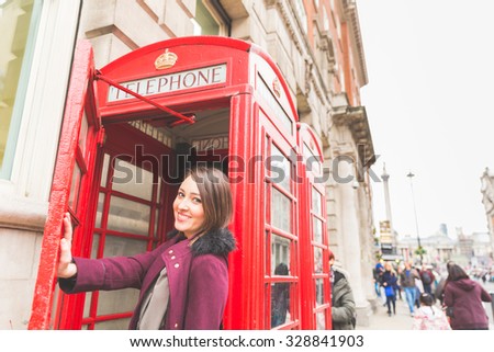Young woman in London in front of a typical red phone booth. She is standing on the entrance, holding the door with her hand, smiling and looking at camera. Tourism and lifestyle concept
