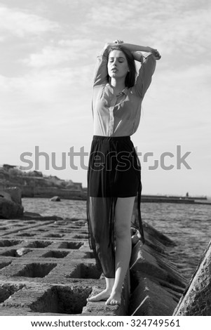 monochrome portrait of a beautiful girl who is dressed in a loose and light clothing and posing near the sea. autumn cold wind ruffling her hair