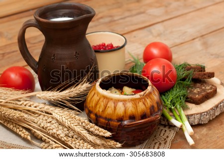 A pottery of cooked vegetables with meat, a crock of milk, a wooden board with a ripe tomato, cucumbers, rye bread and greens on it, and a metal cup of red currants on a wooden surface