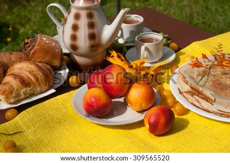 An outdoor composition with tea cups, a tea pot, a plate of pancakes, pastry, ripe fruit and field flowers on a bright yellow and brown table cloth