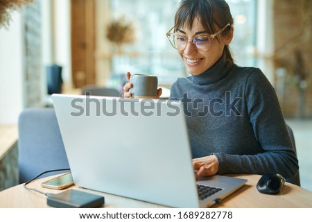 Young smiling business woman in spectacles working on portable laptop computer. Female copywriter sitting at desk and typing on keyboard, working on project, writing down ideas into netbook