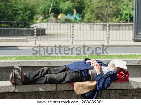 A homeless man is has fallen asleep while reading a bookl on small stone wall. He is using the warmth of the sun to catch up with sleep while searching for food in the night.