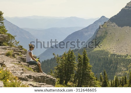 A woman sitting on a rocky ledge looking at a gorgeous view while visiting Glacier National Park in the Rocky Mountains. Large mountain range in the background with copy space