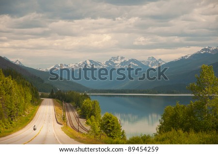 View of highway and majestic snow covered mountains in the distance with a calm lake in the foreground.