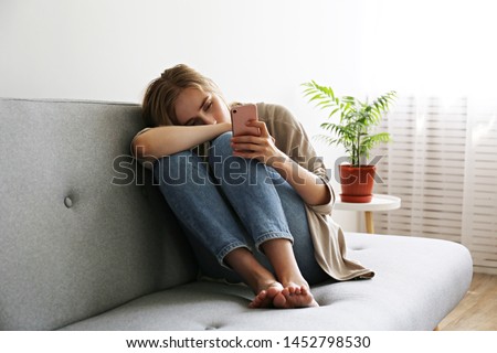 Portrait of beautiful young woman with depressed facial expression sitting on grey textile couch holding her phone. Cyber bullying victim concept. Sad female in her room. Background, copy space. Foto d'archivio © 