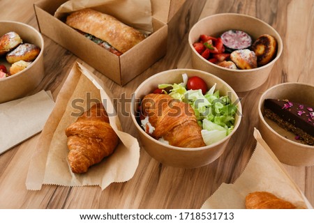 Different takeout food on wooden kitchen table. Italian panini sandwich, french croissant with salmon, strawberry pancakes and chocolate cheesecake. Close up, top view, pov, copy space, background.
