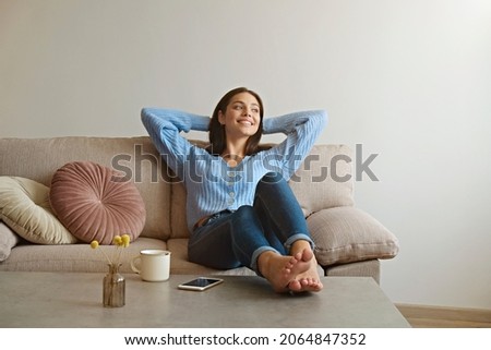 Kick back and relax concept. Young beautiful brunette woman with blissful facial expression alone on the couch with her bare feet on coffee table. Portrait of relaxed female resting at home. Foto d'archivio © 