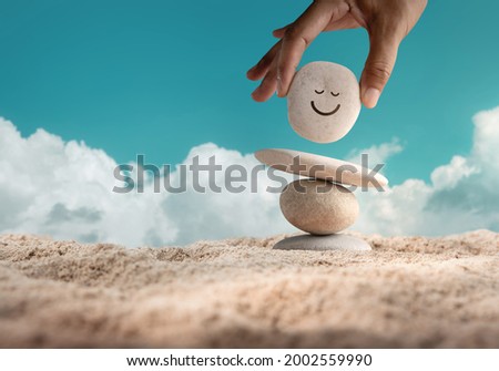 Enjoying Life Concept. Harmony and Positive Mind. Hand Setting Natural Pebble Stone with Smiling Face Cartoon to Balance on Beach Sand. Balancing Body, Mind, Soul and Spirit. Mental Health Practice
