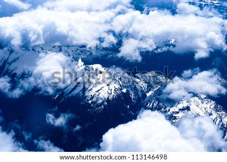 Mountain view from the top through the clouds.   The mountain peaks covered with snow. View from airplane. blue tint