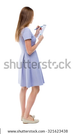 back view of  stands woman takes notes in a notebook. girl  watching. Rear view people collection.    Isolated over white background. The girl in a blue dress stands sideways and draws in a notebook.
