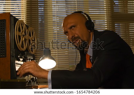 A male spy listens and records conversations on a reel-to-reel tape recorder, intelligence and spying on citizens, and a super-agent with headphones.