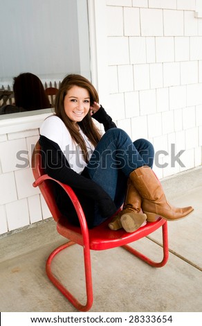 Pretty teen girl sitting in a vintage, red metal chair on the front porch of her vacation beach cabin. The ocean and a picket fence are reflected in the window behind her