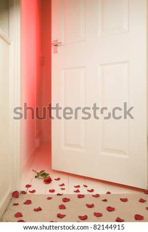 Slightly opened bedroom door with red light coming through and a red rose with rose petals scattered on the carpet floor,