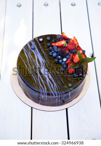 Delicious chocolate mousse cake covered with mirror glaze, decorated with strawberries and blueberries on a white wooden background