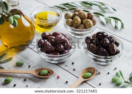 On the right bottle of olive oil to the left 3 bowls with olives, spoons with olives, scattered spices, olive tree branches on wooden white background. 3 different kinds of olives and olive oil.