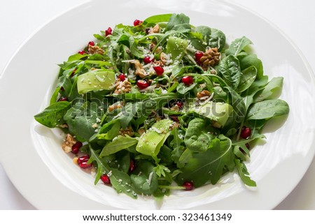 At the center of the frame white plate with a salad of arugula, rocket, avocado, pomegranate seeds, parmesan cheese, spices on a white background. Rocket salad from Greece. Horizontal shot.
